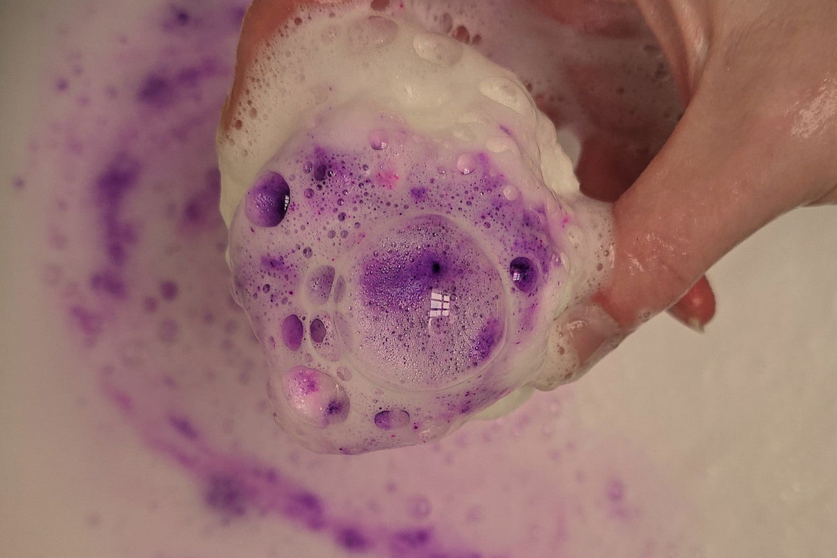 Hand holding a purple bath bomb with bubbles over a bathtub filled with purple water.
